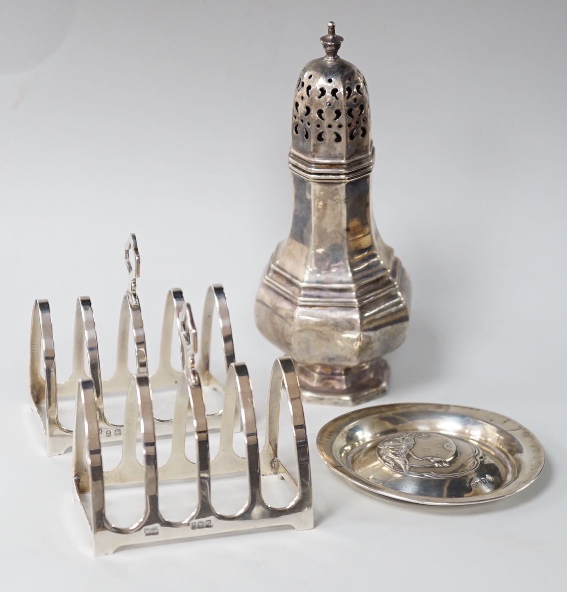 A George V octagonal silver caster, Sheffield 1932, 121 grams, a pair of Walker & Hall toast racks, Birmingham 1921, 101 grams and a trinket dish, decorated with waterlilies, Birmingham 1909, 18 grams